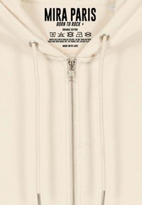 Felpa con zip color crema realizzata in cotone biologico. Presenta una cerniera YKK, coulisse con punte metalliche e istruzioni per la cura stampate.