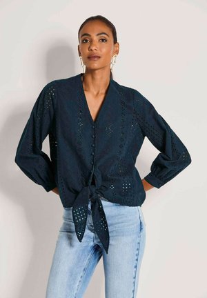 Donkerblauwe blouse met V-hals, lange opgepofte mouwen, eyeletpatroon en een strik in de taille. Gecombineerd met lichtblauwe jeans.