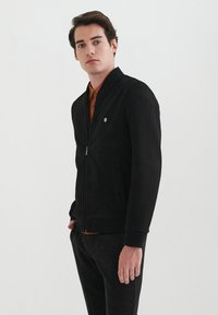 Sort ruskinds bomberjakke med ribbestrikket krave, lynlåslukning og sidelommer. Har et lille emblem på brystet. Båret over en orange skjorte.