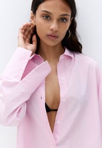 Camicia button-up rosa chiaro con sottili righe verticali bianche e blu, dotata di colletto leggermente aperto e maniche oversize.