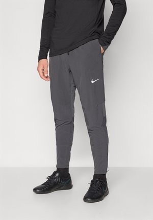 Mężczyzna ubrany w ciemnoszare sportowe spodnie Nike, czarne skarpetki i czarne trampki na białym tle.