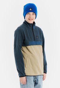 Fleecejacke mit oberem Teil in Marineblau und unterem Teil in Beige, mit Reißverschlusskragen, Seitentasche und blauem "PROTEST"-Logo. Kombiniert mit einer blauen Strickmütze.