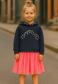 Ein lächelndes junges Mädchen trägt einen dunkelblauen Hoodie mit bunten Sternen und einen leuchtend rosa plissierten Rock. Sie steht vor einem verschwommenen Straßenhintergrund.
