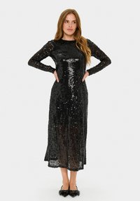 Rochie neagră din paiete, cu mâneci lungi, decolteu rotund și o croială de lungime medie. Materialul are o suprafață strălucitoare și texturată.