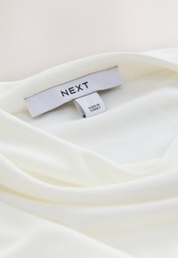 White blouse made of smooth fabric, featuring a draped neckline. Label reads "NEXT," indicating origin from Turkey.