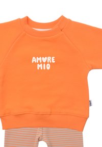 Oranger Pullover mit weißem Text "AMORE MIO" auf der Vorderseite, mit gerippten Bündchen und Saum. Darunter gestreifte hellbraune und beige Leggings.