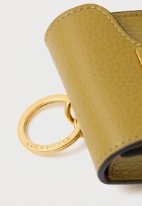 Κίτρινο πορτοφόλι από δέρμα με υφή, με στρογγυλό χρυσό μπρελόκ που φέρει το χαραγμένο όνομα της μάρκας «Coccinelle». Οι άκρες είναι σε σκούρο χρώμα.