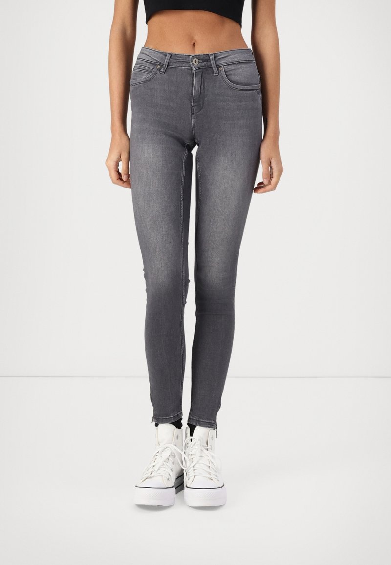 Verwaaide grijze skinny jeans met vijf zakken, een ritssluiting en een lichte rafel bij de enkel, gecombineerd met witte hoge sneakers.