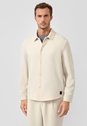 Man draagt een crèmekleurige jas met knopen over een grijs t-shirt en bijpassende crèmekleurige broek, staand tegen een lichtgrijze achtergrond.