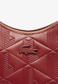 Sac en cuir rouge texturé arborant un motif géométrique, une fermeture éclair dorée et un logo crocodile en relief bien en vue.