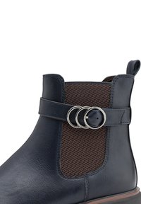 Bottine en cuir bleu foncé avec un panneau textile à motifs marron, une sangle décorative avec deux anneaux en métal argenté, une surface texturée et une languette d'enfilage.