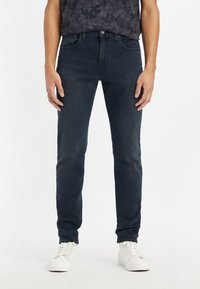 Vaqueros de mezclilla oscura con un ajuste slim, que cuentan con el clásico estilo de cinco bolsillos y una textura suave. Combinados con zapatillas blancas.