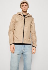 Beige lightweight jacket with a hood, front zip closure, elastic cuffs, and black trim, worn over a black shirt and light blue jeans.
