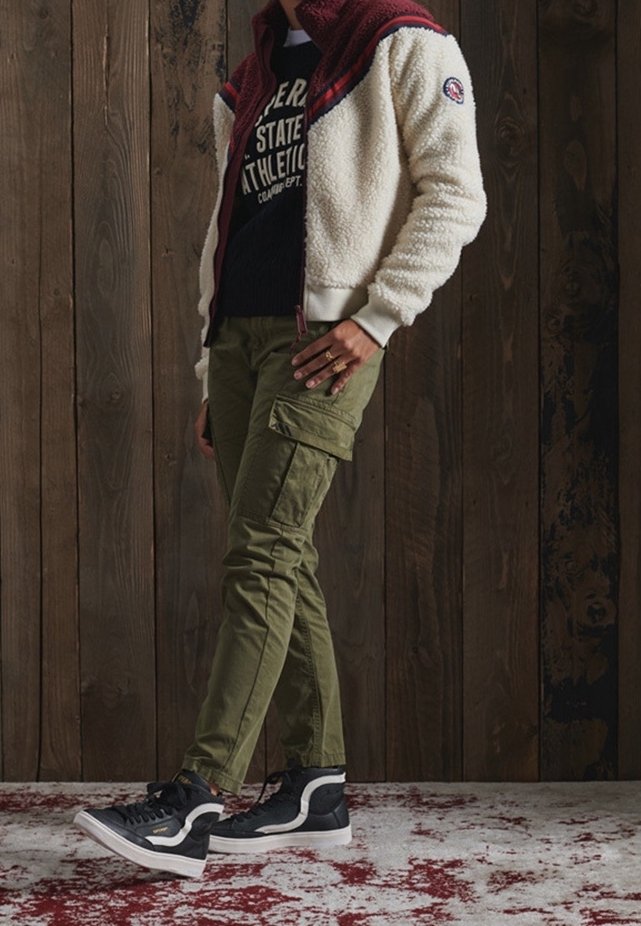 Personne portant un pantalon cargo olive, des baskets noires et blanches, un t-shirt graphique noir et une veste polaire blanche avec une capuche bordeaux devant un mur en bois.