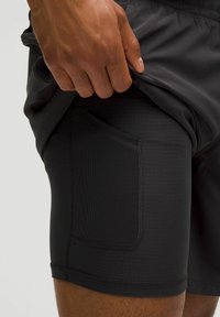 Rukou zdvíhajúce vonkajšie čierne šortky, odhaľujúce vnútorné čierne kompresné šortky s bočnou sieťovinovou kapsou na stehne.