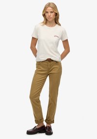 Camiseta de color gris claro con logo rojo, combinada con pantalones beige de corte slim. Zapatos marrones tipo slip-on, con los puños enrollados en los tobillos.