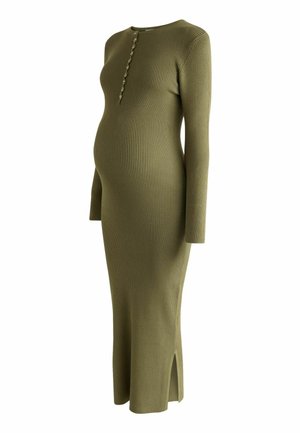 Pregnant woman standing in an olive green ribbed long-sleeve dress and beige sneakers against a plain beige background.