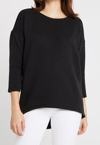 Musta pitkähihainen toppi, jossa on pyöreä pääntie, roikkuvat olkapäät ja kaareva helma. Kankaan pinnassa on hienovarainen teksturoitu kuvio.