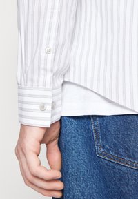 Bílá košile s dlouhými rukávy a šedými pruhy, s knoflíkovými manžetami, spárovaná s modrými džíny. Viditelné detaily textury a šití.