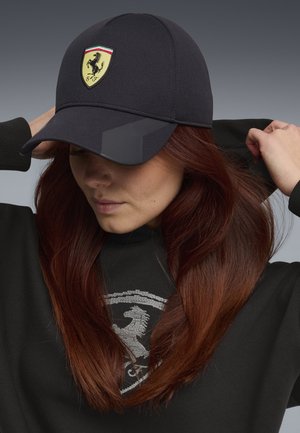 Vrouw met lang rood haar die een zwarte pet draagt met een geel Ferrari-logo en een zwarte hoodie met een bijpassend embleem, terwijl ze haar capuchon rechtzet.