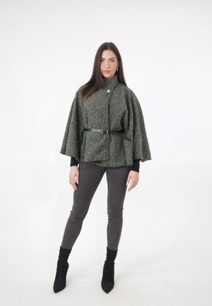 Vrouw draagt een gestructureerde grijze cape met ceintuur, donkere aansluitende broek en zwarte enkellaarsjes, staande tegen een witte achtergrond.