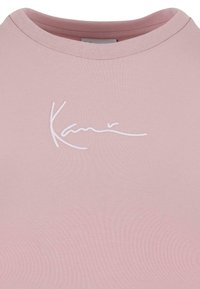 Lyserød t-shirt med rund halsudskæring og et hvidt broderet signaturdesign på forsiden. Strækbart, ribbet stof.