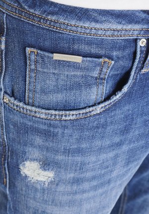 Nahaufnahme einer Tasche einer blauen Jeans aus Denim mit sichtbaren orangefarbenen Nähten, Metallnieten, kleinem Markenetikett und einem verblassten, abgenutzten Bereich unter der Tasche.