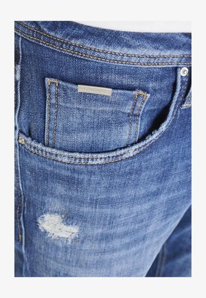 Nahaufnahme einer Tasche einer blauen Jeans aus Denim mit sichtbaren orangefarbenen Nähten, Metallnieten, kleinem Markenetikett und einem verblassten, abgenutzten Bereich unter der Tasche.