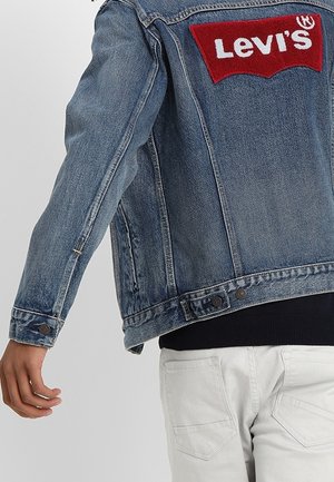 Person wearing a blue denim Levi's jacket with a large red and white logo on the back, paired with white pants.
