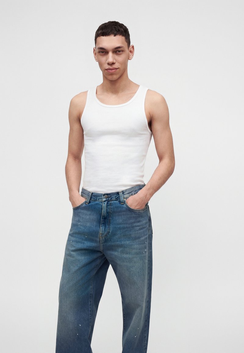 Jeune homme aux cheveux courts et foncés portant un débardeur blanc et un jean bleu ample, debout les mains dans les poches devant un fond uni.