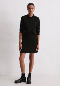 Cardigan noir à boutons avec un col, associé à une jupe noire moulante et des chaussures noires chunky, avec des chaussettes de cheville côtelées.