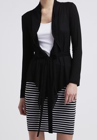 Černý zavinovací kardigán s volným designem a dlouhými rukávy, kombinovaný s upnutou pruhovanou sukní ke kolenům v černé a bílé barvě.