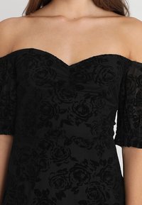 Vestido negro off-the-shoulder con tejido texturizado y patrones de rosas, un torso ajustado y mangas cortas abullonadas.