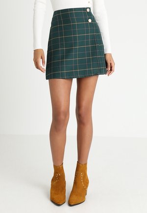 Green plaid mini skirt with tan accents, featuring two decorative buttons at the waist. Paired with mustard suede ankle boots with studs.