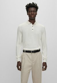 Langärmliges, elfenbeinfarbenes Poloshirt mit gestrickter Textur, drei schwarze Knöpfe am Kragen, kombiniert mit hellbeigen, maßgeschneiderten Hosen und einem schwarzen Gürtel.