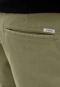 Κοντινή λήψη της πίσω τσέπης σε χακί παντελόνι με μικρή λευκή ετικέτα που γράφει «JJ REBEL» ραμμένη πάνω από τη γραμμή της τσέπης.