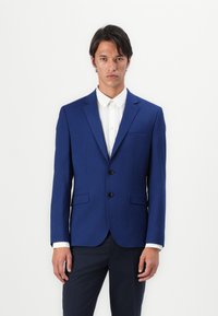 Blå blazer laget av glatt stoff, med to svarte knapper, klappet krage, sidelommer og en skreddersydd passform, båret over en hvit skjorte.
