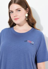 Niebieska koszulka z krótkim rękawem wykonana z miękkiego materiału, z napisem "OPTIMIST by PURE" w kontrastowych kolorach na piersi.