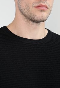 Pull noir avec un motif texturé, col rond et bordures côtelées. Vue en gros plan mettant en valeur les détails du tissu et du col.