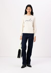 Pull créme avec "voilà" en script noir, associé à un jean évasé bleu marine et un sac à main blanc texturé ; des chaussures à talons noires complètent le look.
