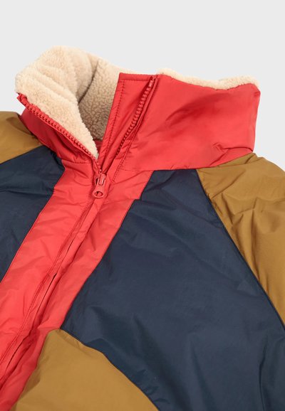 Giacca puffer a blocchi di colore con pannelli rossi, blu navy e verde oliva. Ha una chiusura con zip e un colletto morbido foderato in fleece beige.