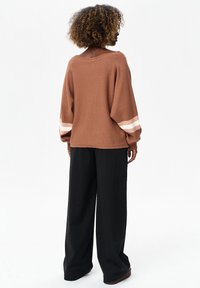 Vrouw met krullend haar, een losse bruine trui met witte strepen op de mouwen en brede zwarte broek, staand met de rug naar de kijker.