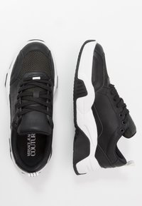 Black and white Versace Jeans Couture sneakers with mesh and leather uppers, visible logo, black laces, and chunky soles shown from top and side.