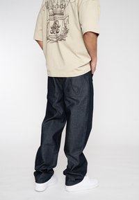T-shirt beige surdimensionnée avec un design graphique sombre au dos, associé à un pantalon en denim bleu foncé, ample et à des baskets blanches.
