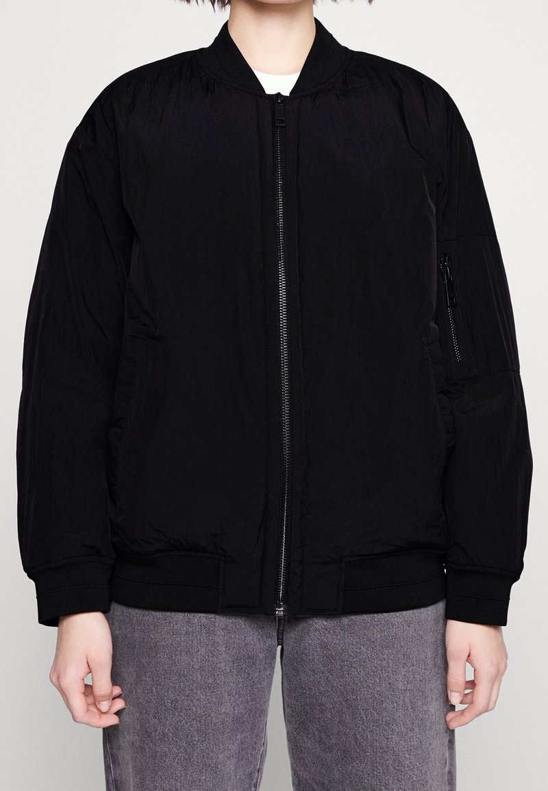 Veste bomber noire avec fermeture éclair à l'avant, poignets côtelés et ourlet. Comprend des poches latérales et une texture lisse, fabriquée en matériau léger.