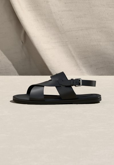 Μαύρο δερμάτινο σανδάλι με ανοιχτά δάχτυλα, ευρείς σταυρωτοί ιμάντες και ασημένια πόρπη, σε ανοιχτή επιφάνεια με φόντο σκιασμένο μπεζ ύφασμα.