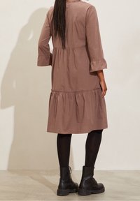 Femme avec de longs cheveux tressés portant une robe midi mauve à jupe à volants, des bas noirs et des bottines à lacets noires, se tenant sur un sol clair.