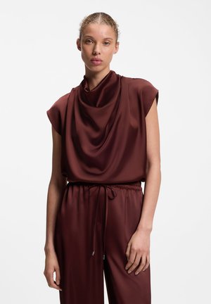 Femme à la peau claire et cheveux attachés portant un haut en satin marron foncé sans manches et un pantalon assorti avec une taille à cordon.