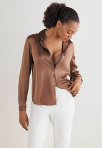 Bruine satijnen blouse met lange mouwen, kraag, knoopsluiting aan de voorkant en zijsplitten. Gemonteerd over een kanten bh, gecombineerd met een witte pantalon.