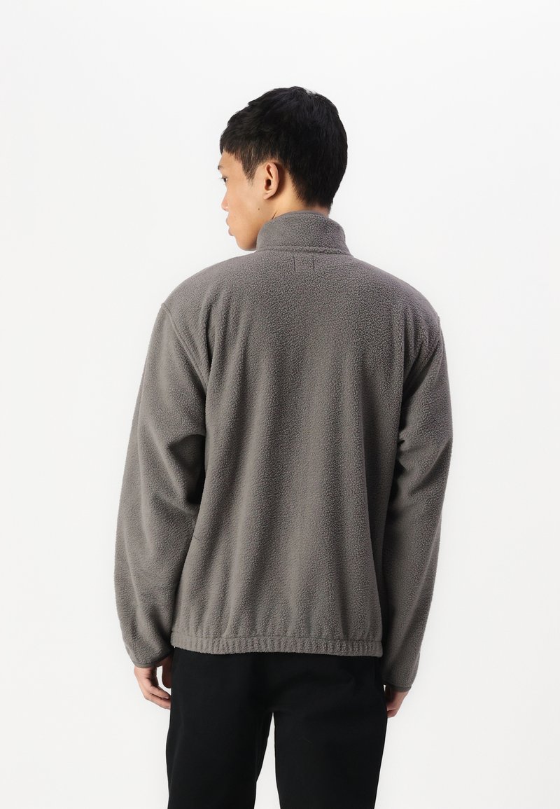 Graue Fleecejacke mit hohem Kragen, langen Ärmeln, elastischem Saum und strukturiertem Stoff. Die Rückseite hat subtile Nähte.